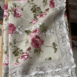 Puede incluir: Un mantel de lino beige con un patrón floral de rosas rosas y hojas verdes. El mantel tiene un borde de encaje blanco.