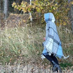 Könnte beinhalten: Eine Person trägt einen Kapuzenponcho in blau-weißem Karomuster mit Fransenbesatz und geht durch ein Grasfeld mit Herbstlaub im Hintergrund.