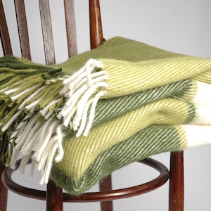 Könnte beinhalten: Eine grün-weiß gestreifte Wolldecke mit Fransen auf einem Holzstuhl. Die Decke ist in der Mitte gefaltet und hat eine weiche, flauschige Textur.