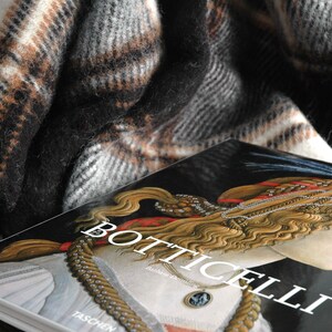 May include: A close-up of a book titled "Botticelli" featuring a painting of a woman with long, flowing hair and a gold necklace. The book is partially covered by a brown and black checked blanket.