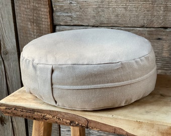 May include: A round, beige meditation cushion made of linen fabric. The cushion is sitting on a wooden surface.