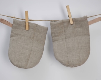 May include: Two beige linen oven mitts hanging from a clothesline with wooden clothespins. Each mitt has a loop for hanging and a quilted design. The background is a plain white wall.