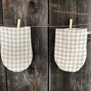 Puede incluir: Dos manoplas de horno de vichy beige y blanco colgadas sobre un fondo de madera.