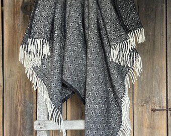 Puede incluir: Manta plegada con estampado en blanco y negro con flecos blancos. La manta está colocada sobre una escalera de madera contra una pared de madera desgastada. El patrón es un diseño de diamantes repetido.