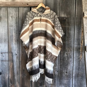 Puede incluir: Un poncho a rayas marrón, blanco y beige con capucha y flecos. El poncho está colgado en una pared de madera.