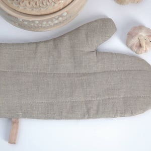 Può includere: Un guanto da forno in lino beige con bordino rosso e un cinturino beige. Il guanto è trapuntato e ha una superficie testurizzata.