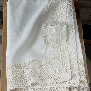Puede incluir: Mantel de lino blanco con un delicado borde de encaje. El encaje es un patrón intrincado de pequeñas flores y hojas. El mantel está doblado por la mitad y está sobre una superficie de madera.