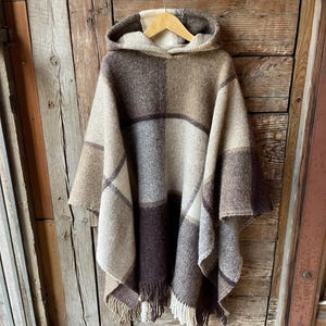 Op de afbeelding: Een bruine en beige poncho met capuchon met een ruitpatroon. De poncho heeft een franje rand en hangt aan een houten hanger.