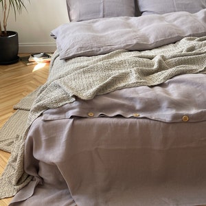 Op de afbeelding: Een licht lavendelgrijze dekbedovertrekset met een knoopsluiting van ronde houten knopen. Het beddengoed is bedekt met een gestructureerde, lichtbeige wafeldekentje dat eroverheen gedrapeerd is, samen met twee bijpassende kussenslopen.