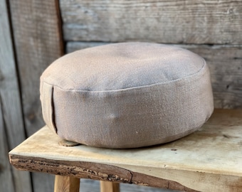May include: A round, light brown meditation cushion with a textured fabric cover. The cushion is sitting on a wooden surface.