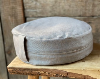 May include: A round gray meditation cushion with a fabric cover and a sewn seam around the edge. The cushion is sitting on a wooden surface.
