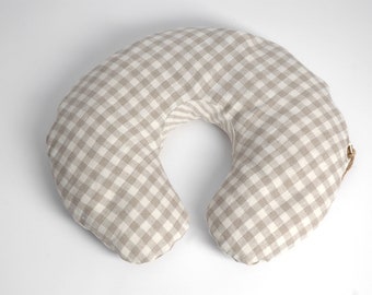 May include: A beige and white gingham patterned nursing pillow. The pillow is shaped like a crescent moon and has a brown tie.