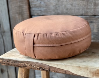 May include: A round, terracotta-colored meditation cushion with a simple design and a zipper closure. The cushion is sitting on a wooden surface.