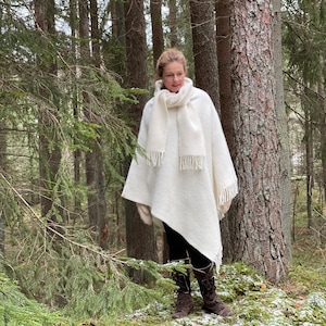 Può includere: Un poncho bianco lavorato a maglia con dettagli a frangia indossato da una persona in un ambiente boschivo. Il poncho è drappeggiato sulle spalle della persona e ha un design lungo e fluido.