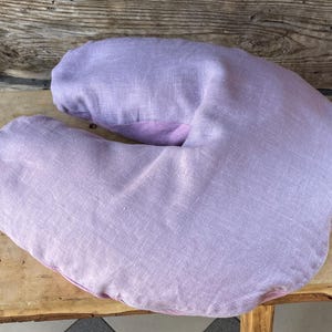 May include: A lavender-colored linen bolster pillow shaped like a crescent moon. The pillow is made of soft, natural linen and is perfect for supporting the neck and head during massage or relaxation.