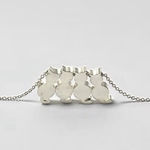 May include: A silver-colored necklace featuring four cat silhouettes in a row. The cats are connected and suspended from a delicate chain. The necklace is displayed against a light gray background.