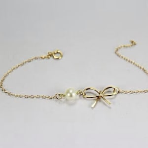 Puede incluir: Una delicada pulsera dorada con un dije de lazo y una sola perla. La pulsera tiene una cadena fina y un cierre de broche. El dije de lazo está centrado en la pulsera.