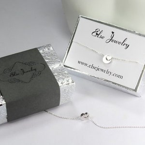 Puede incluir: Un collar de plata con un peque&ntilde;o colgante circular, empaquetado en una caja gris con el texto "Else Jewelry" y una caja blanca con el texto "Else Jewelry" y la direcci&oacute;n del sitio web "www.elsejewelry.com".