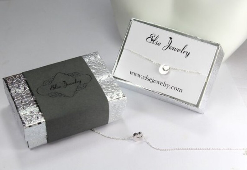Puede incluir: Un collar de plata con un peque&ntilde;o colgante circular, empaquetado en una caja gris con el texto "Else Jewelry" y una caja blanca con el texto "Else Jewelry" y la direcci&oacute;n del sitio web "www.elsejewelry.com".