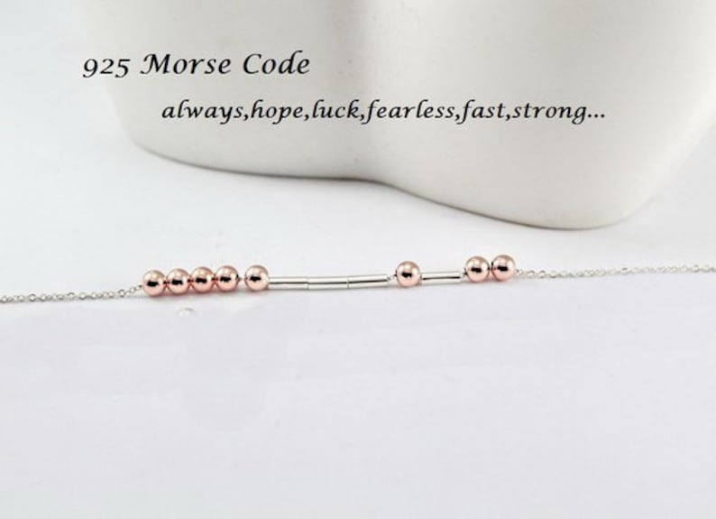 Pu&ograve; includere: Un braccialetto a catena d'argento con una barra d'argento e perline in oro rosa. Il braccialetto ha il testo "925 Morse Code" e "always, hope, luck, fearless, fast, strong..."