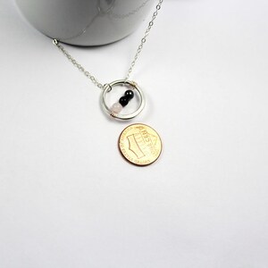 May include: A silver chain necklace with a circle pendant featuring three small beads: one pink, one black, and one black. The necklace is displayed on a white surface next to a US penny.
