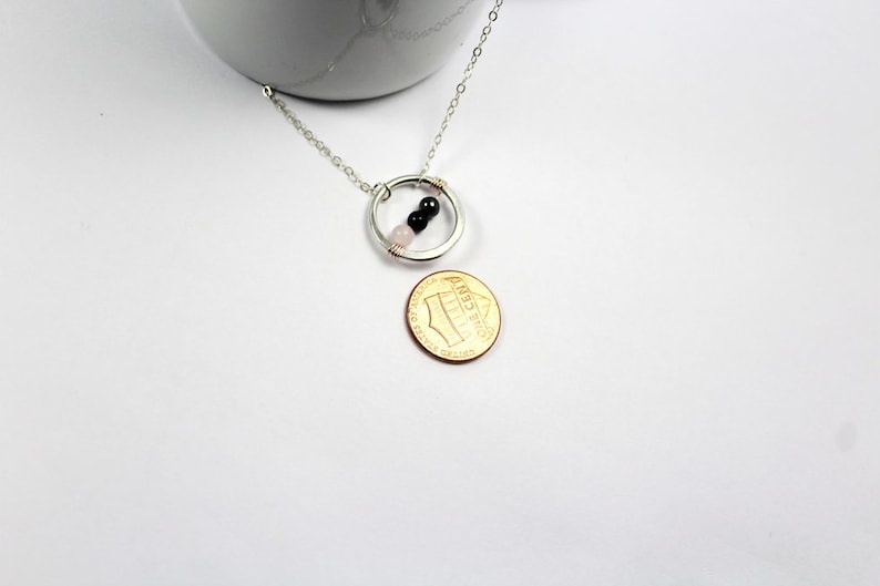 May include: A silver chain necklace with a circle pendant featuring three small beads: one pink, one black, and one black. The necklace is displayed on a white surface next to a US penny.