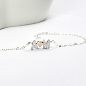 May include: A silver chain bracelet with two silver letters, "A" and "M", and a small rose gold heart charm in between.