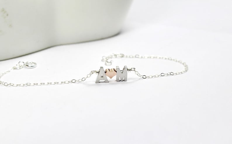 May include: A silver chain bracelet with two silver letters, "A" and "M", and a small rose gold heart charm in between.