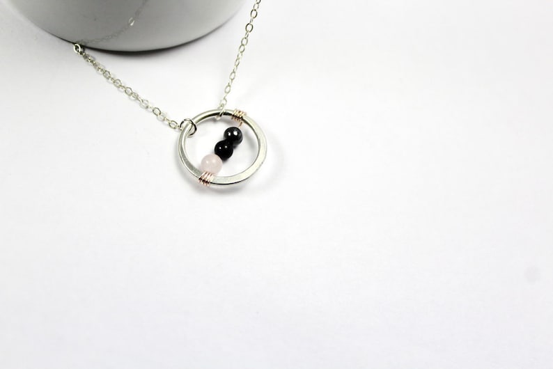 May include: A silver necklace with a circle pendant featuring three beads: a pink, a black, and another black bead.