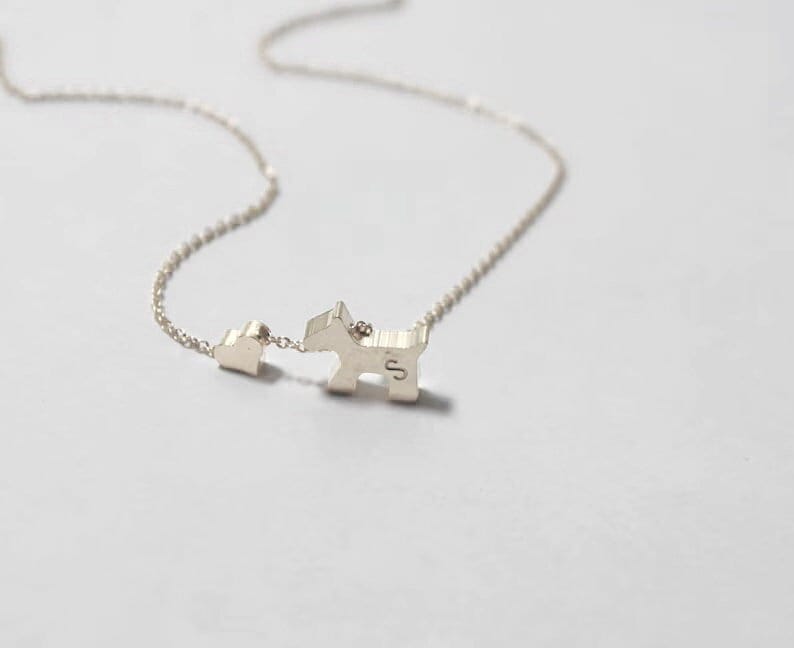 May include: A silver necklace with a small heart charm and a larger charm shaped like a dog. The dog charm has the letter "S" engraved on it.