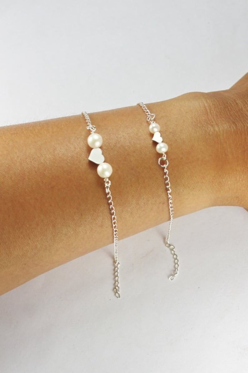 K&ouml;nnte beinhalten: Zwei silberne Kettenarmb&auml;nder mit kleinen wei&szlig;en Perlen und silbernen Herzanh&auml;ngern.