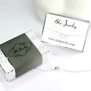 Pu&ograve; includere: Una collana d'argento con un piccolo ciondolo a forma di cuore, confezionata in una scatola grigia con la scritta "Else Jewelry" su di essa. La scatola &egrave; appoggiata su una superficie bianca. La collana &egrave; anche mostrata su una carta bianca con la scritta "Else Jewelry" e "www.elsejewelry.com".