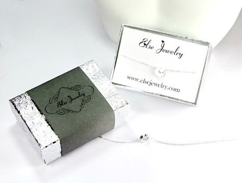 Pu&ograve; includere: Una collana d'argento con un piccolo ciondolo a forma di cuore, confezionata in una scatola grigia con la scritta "Else Jewelry" su di essa. La scatola &egrave; appoggiata su una superficie bianca. La collana &egrave; anche mostrata su una carta bianca con la scritta "Else Jewelry" e "www.elsejewelry.com".