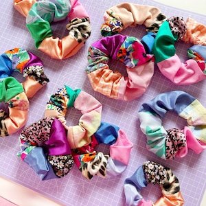 Peut inclure: Une collection de chouchous en tissu colorés avec différents motifs, notamment des motifs floraux, animaliers et géométriques. Les chouchous sont disposés sur une surface quadrillée violette.