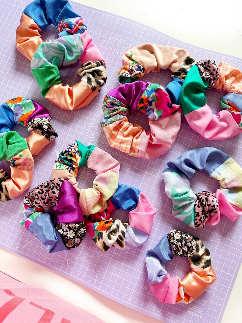 Op de afbeelding: Een verzameling kleurrijke stoffen scrunchies met verschillende patronen, waaronder bloemen, dierenprint en geometrische ontwerpen. De scrunchies zijn gerangschikt op een paarse geruite ondergrond.