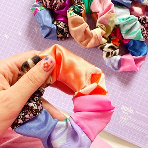 Op de afbeelding: Een kleurrijke haarband gemaakt van meerdere stofresten die aan elkaar zijn genaaid. De haarband is in een hand, en de stofresten omvatten roze, oranje, blauw, paars, groen en wit.