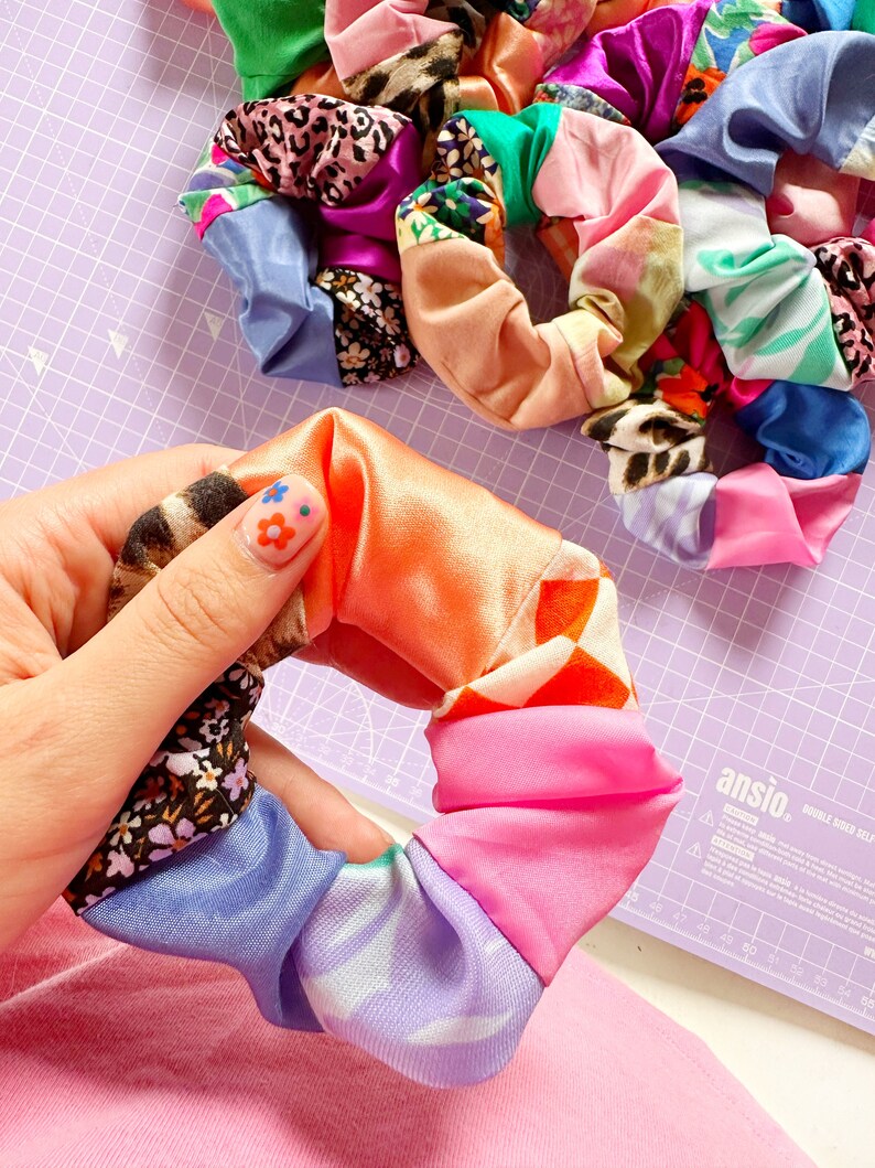 Op de afbeelding: Een kleurrijke haarband gemaakt van meerdere stofresten die aan elkaar zijn genaaid. De haarband is in een hand, en de stofresten omvatten roze, oranje, blauw, paars, groen en wit.