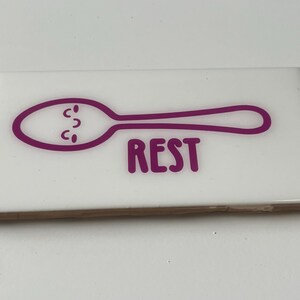 May include: White ceramic tile with a purple outline of a spoon with a face and the word "REST" underneath.