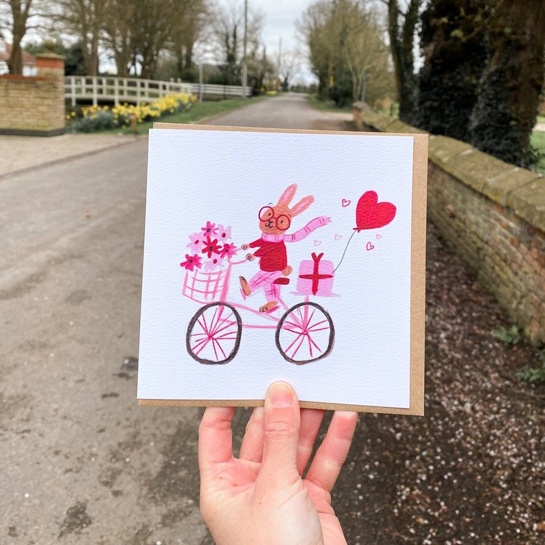 May include: A pink card with a watercolor illustration of a bunny wearing glasses and a scarf riding a pink bicycle with a basket of flowers and a pink gift box. The bunny is holding a pink heart balloon.