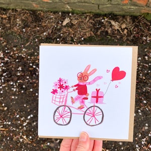 May include: A pink and white illustrated greeting card featuring a bunny wearing glasses and a pink scarf riding a pink bicycle with a basket of pink flowers and a pink gift box. The bunny is holding a pink heart balloon.