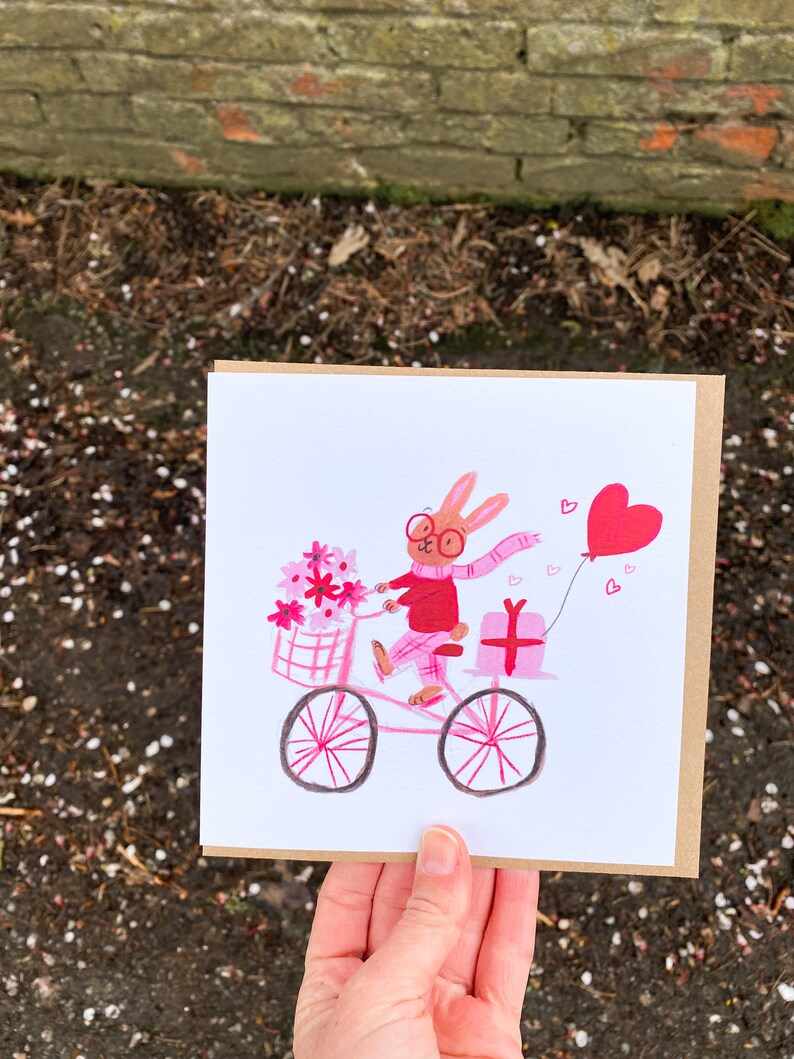 May include: A pink and white illustrated greeting card featuring a bunny wearing glasses and a pink scarf riding a pink bicycle with a basket of pink flowers and a pink gift box. The bunny is holding a pink heart balloon.