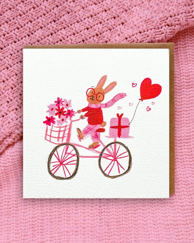 Illustrated card featuring a cheerful bunny riding a pink bicycle, carrying flowers, a gift, and a heart-shaped balloon. The bunny wears glasses, a red sweater, and a pink scarf. Perfect for Valentine's Day or any occasion to share love and joy.