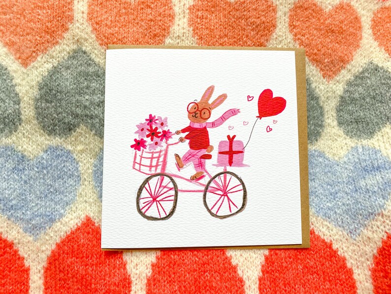May include: A pink bicycle with a basket of pink flowers, a pink gift box, and a pink heart balloon. A brown rabbit with glasses and a red scarf is riding the bicycle.