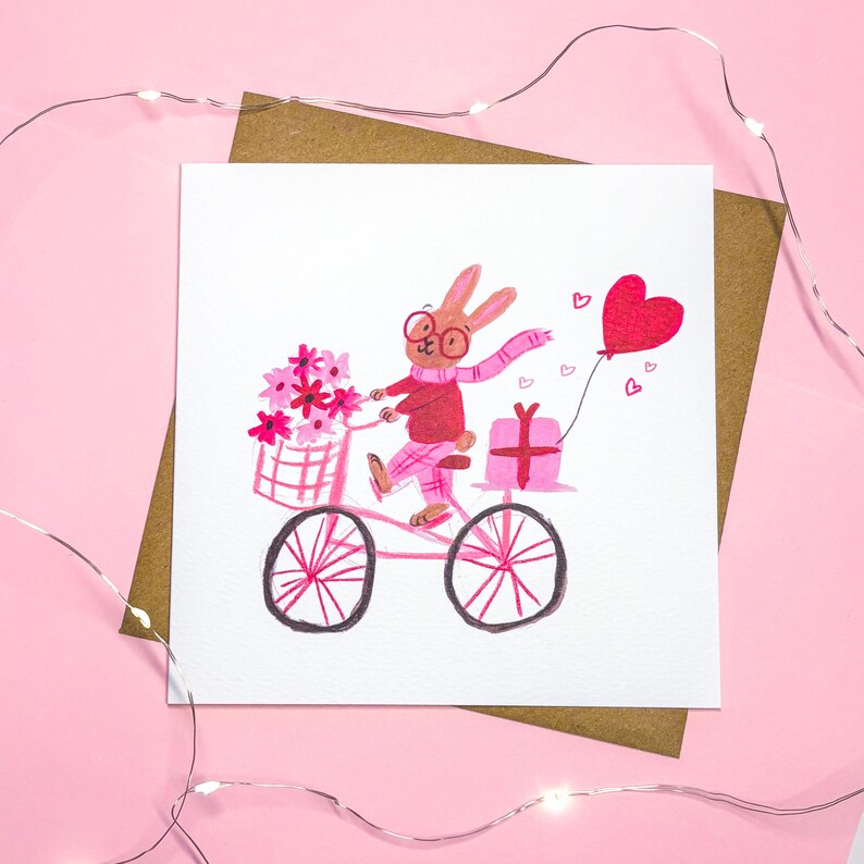May include: A pink and red illustrated card featuring a bunny wearing glasses and a pink scarf riding a pink bicycle with a basket of pink flowers. The bunny is carrying a pink gift box and a red heart balloon.