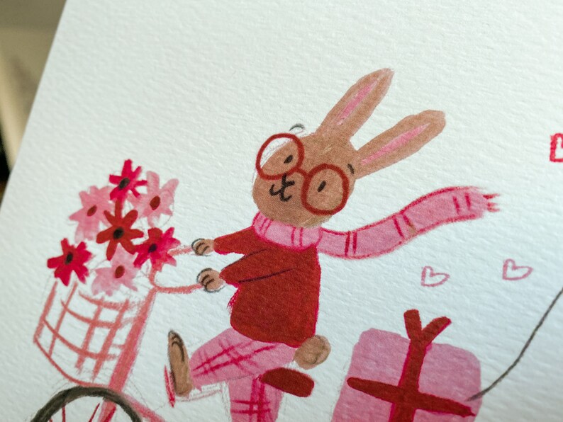 May include: A brown rabbit wearing red glasses, a red sweater, and a pink scarf rides a pink bicycle with a basket of pink and red flowers. The rabbit is carrying a pink gift box.