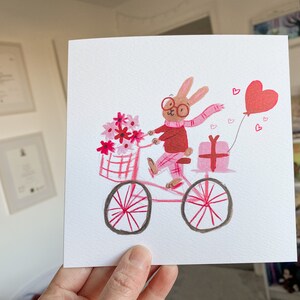 May include: A pink bicycle with a basket full of pink flowers, a pink gift box, and a red heart balloon. A brown rabbit wearing glasses and a pink scarf is riding the bicycle.