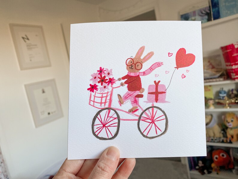 May include: A pink bicycle with a basket full of pink flowers, a pink gift box, and a red heart balloon. A brown rabbit wearing glasses and a pink scarf is riding the bicycle.