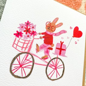 May include: A pink bicycle with a basket full of pink flowers is being ridden by a brown rabbit wearing red glasses, a red sweater, and a pink scarf. The rabbit is carrying a pink gift box and a pink heart balloon.