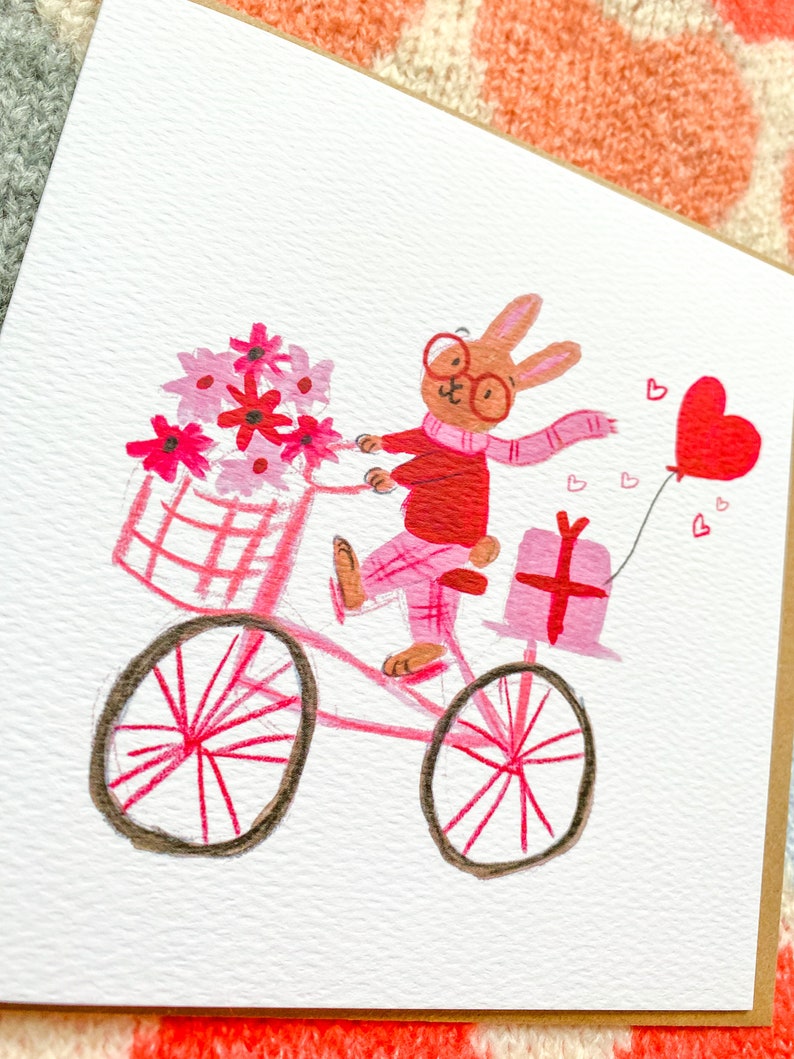 May include: A pink bicycle with a basket full of pink flowers is being ridden by a brown rabbit wearing red glasses, a red sweater, and a pink scarf. The rabbit is carrying a pink gift box and a pink heart balloon.