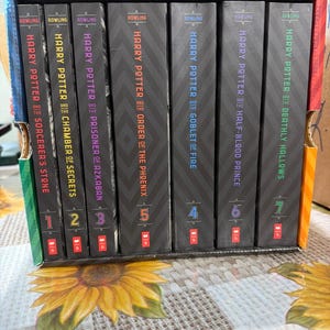 May include: A seven-volume Harry Potter book set housed in a black box with a chevron pattern. The spines feature colorful text and numbers one through seven, including titles like Sorcerer's Stone, Chamber of Secrets, and Deathly Hallows by Rowling.